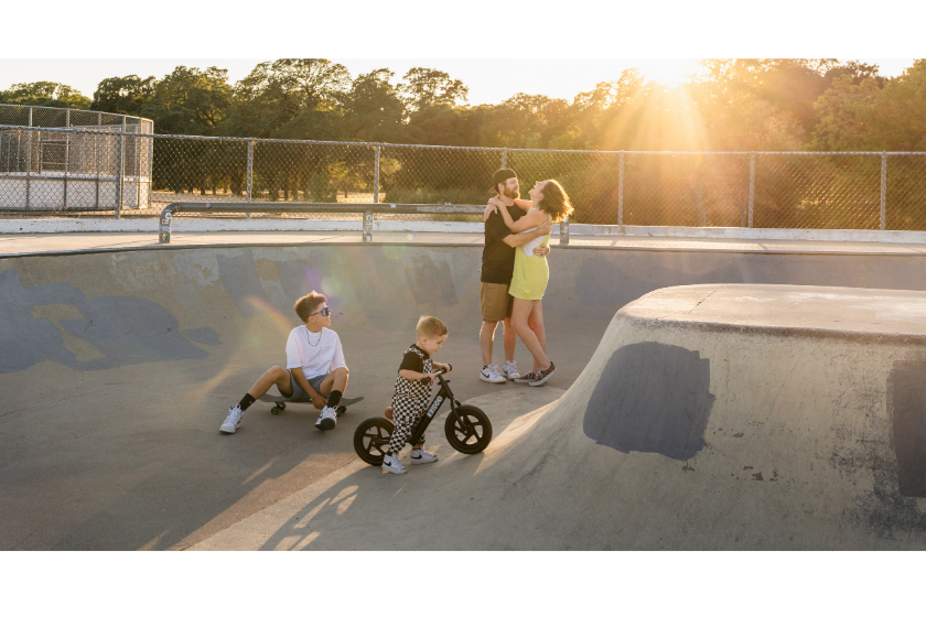 Rocklin family portrait session at skatepark during golden hour, parents embracing while kids ride bikes around them.